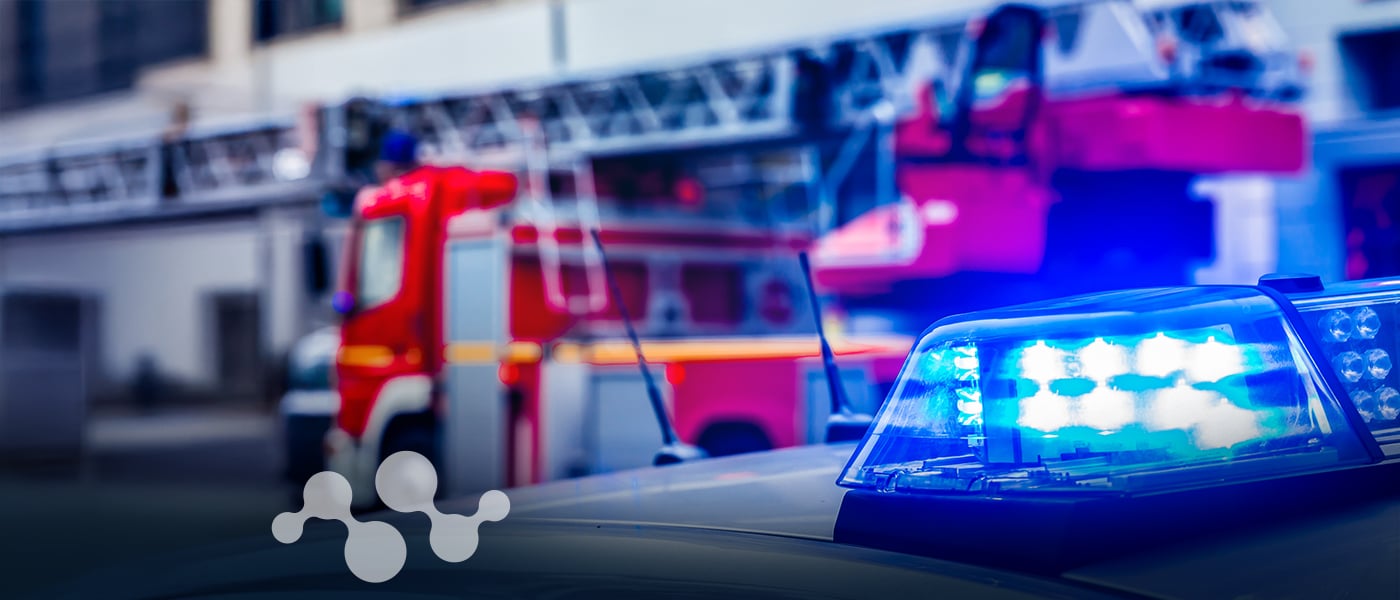 Police vehicle with illuminated blue emergency lights in the foreground and a red fire truck in the background, representing first responders who rely on FirstNet® certified communication devices for emergency operations.