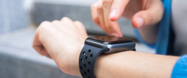 The image shows a close-up of a person's wrist wearing a black smartwatch or fitness tracker with a perforated sport band. A finger from their other hand is touching or interacting with the watch display. The background is blurred, giving the image a soft focus that emphasizes the smartwatch interaction. The image appears to be showcasing how someone would use the touchscreen functionality of the wearable device in everyday use.